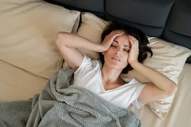 Femme allongée dans son lit, mains sur le visage, illustrant les troubles du sommeil et leurs effets