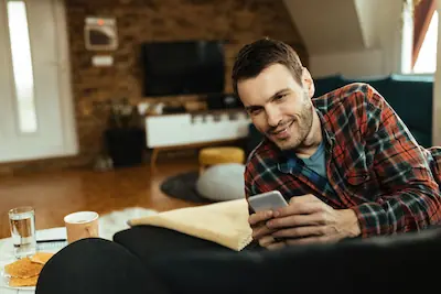 Homme souriant sur son canapé consultant son téléphone, illustrant la facilité d'achat d'un bon cadeau massage en ligne