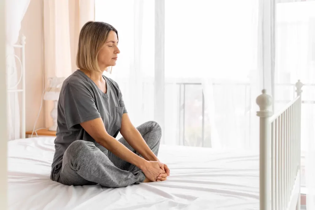 Femme assise les yeux fermes sur son lit dans la lumiere du matin, entre fatigue mentale et moment de recentrage