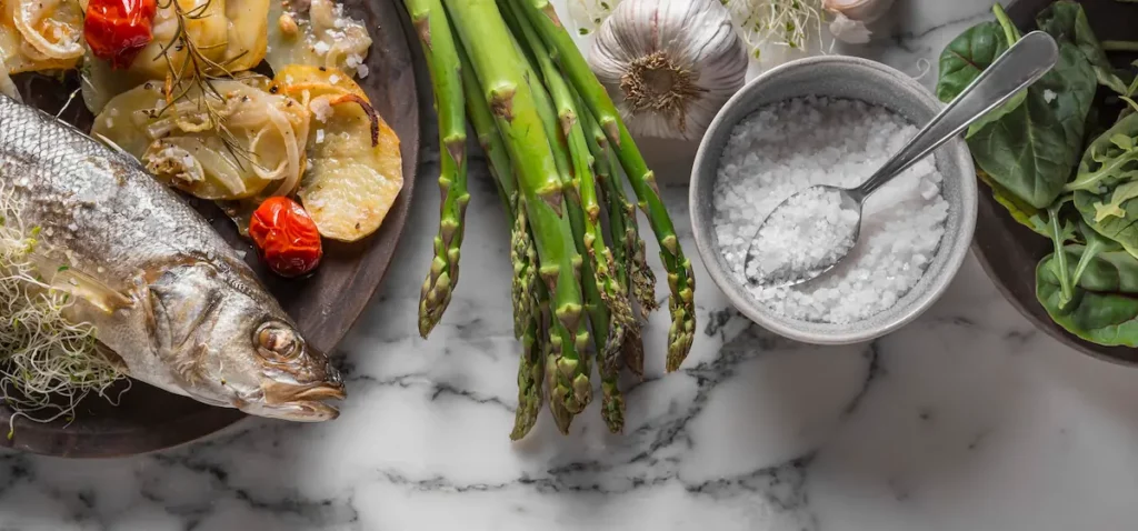 Flat-lay alimentaire : poisson cuit, asperges vertes, ail, épinards et bol de sel — base d'une alimentation anti-inflammatoire pour la thyroïde