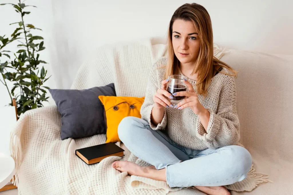 Femme assise en tailleur sur un canapé, tasse dans les mains, expression pensive — fatigue chronique liée à l'hypothyroïdie
