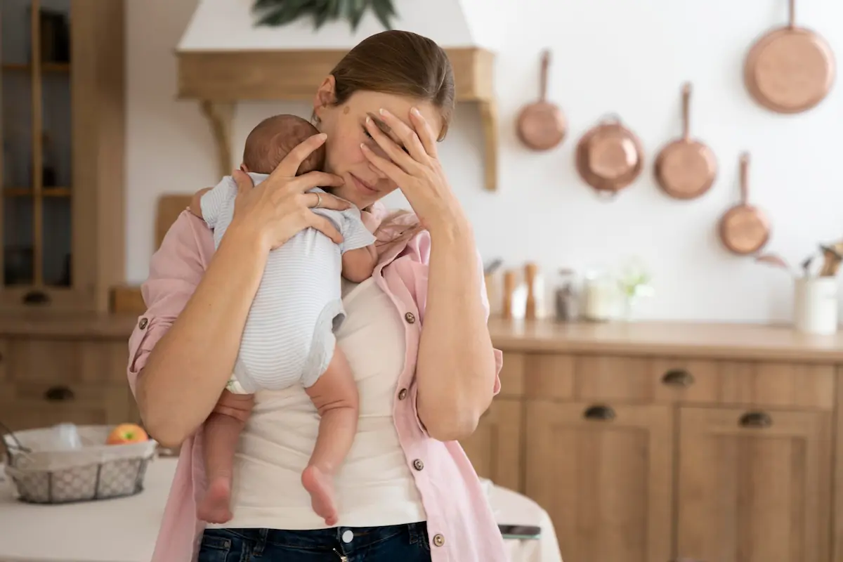 Jeune maman épuisée portant son bébé dans la cuisine, main sur le visage, stress parental.