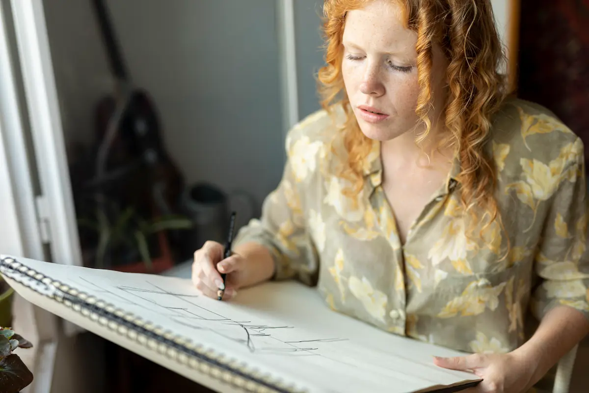 epuisement-creatif-femme-rousse-dessin-fenetre Jeune femme rousse pensive dessinant dans un carnet à spirales près d'une fenêtre, ambiance contemplative