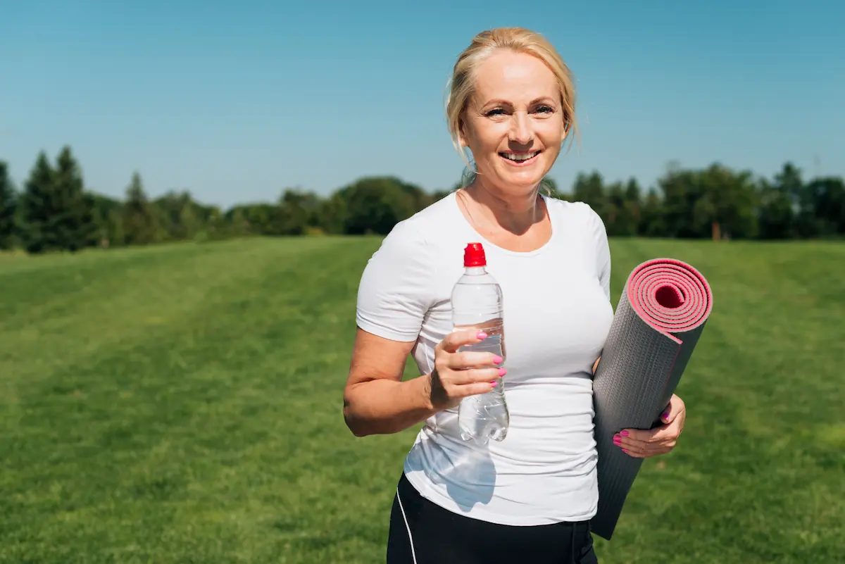 perte-poids-45-ans-femme-yoga-vitalite Femme d'une cinquantaine d'années souriante avec tapis de yoga et bouteille d'eau dans un champ verdoyant