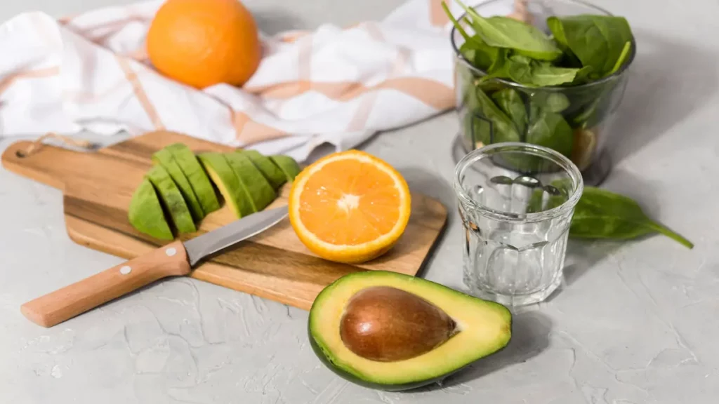 Demi-avocat, orange coupée et épinards frais près d’un verre d’eau, sur un plan de travail clair