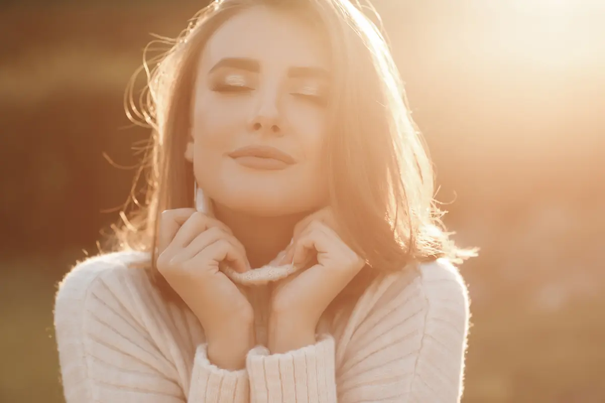 symbolique-de-la-peau-femme-apaisee-soleil Femme aux yeux fermés, visage apaisé sous une lumière dorée, mains repliées près du col d’un pull clair