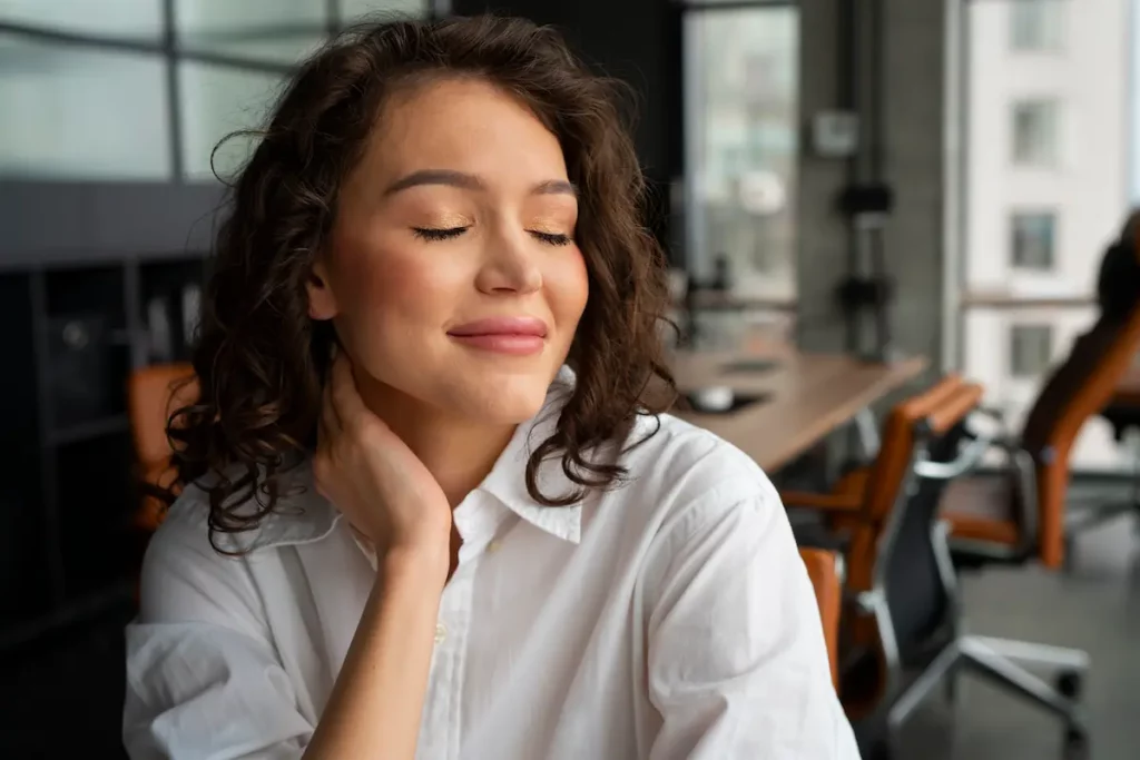 Femme souriante aux yeux clos, main posée sur la nuque lors d’une pause détente dans un bureau